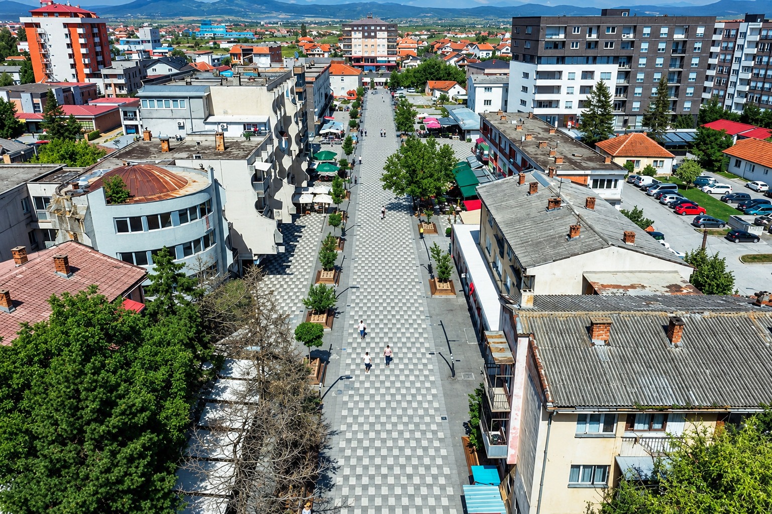 Aerial view of the pedestrian square in Lipjan, paved promenade lined with trees and cafés between the surrounding apartment blocks