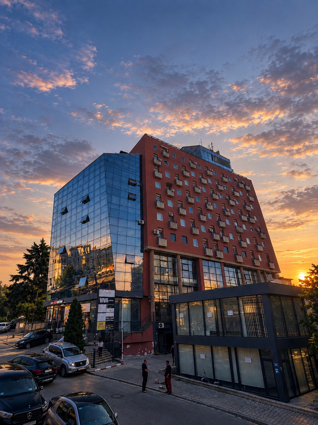 Kompleksi Donika 1's building at sunset, glass and brick facade against a colourful sky